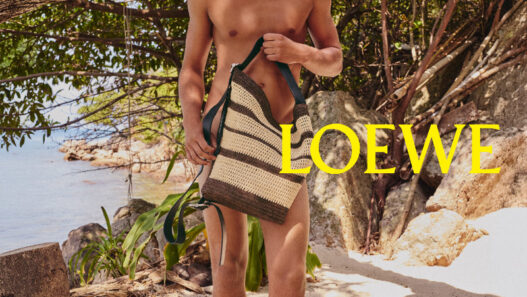 Male model standing on a sunny beach under a tree, shirtless, holding a striped woven tote bag; LOEWE logo in yellow across the image.