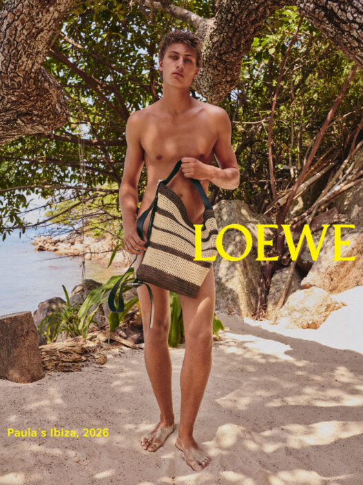 Male model standing on a sunny beach under a tree, shirtless, holding a striped woven tote bag; LOEWE logo in yellow across the image.