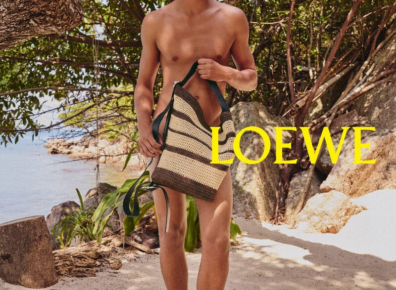 Male model standing on a sunny beach under a tree, shirtless, holding a striped woven tote bag; LOEWE logo in yellow across the image.