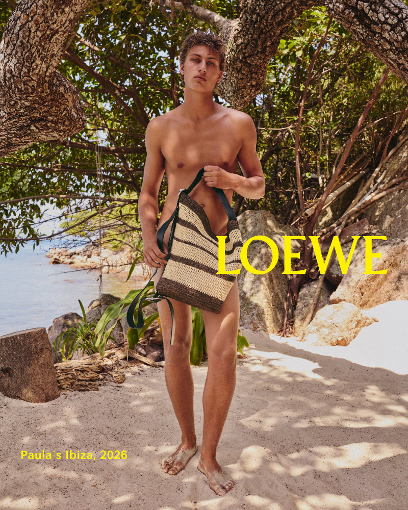 Male model standing on a sunny beach under a tree, shirtless, holding a striped woven tote bag; LOEWE logo in yellow across the image.