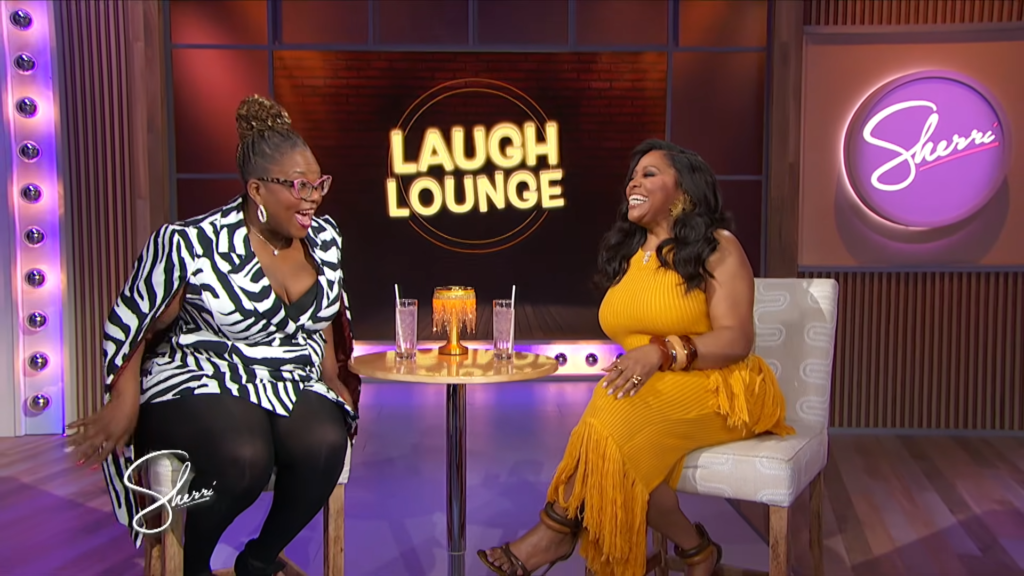 Two women sit on a TV talk-show set, laughing together at a small round table with two drinks and a golden centerpiece behind them (Laugh Lounge sign visible).