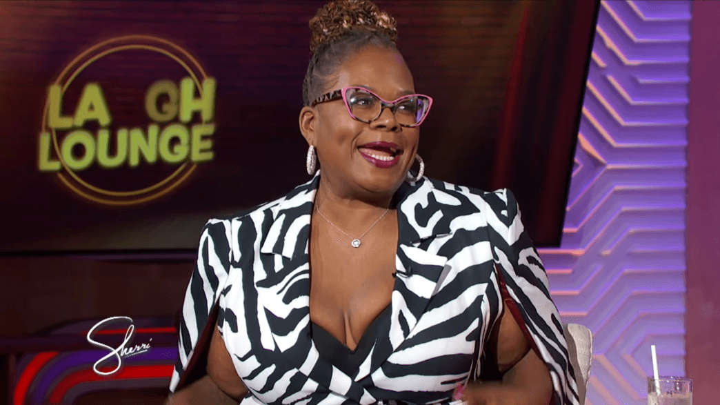 Woman guest wearing a black-and-white zebra print blazer with pink glasses and large hoops on a TV talk show set.