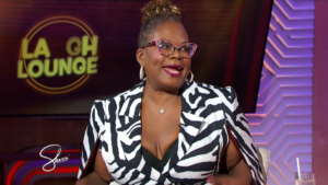 Woman guest wearing a black-and-white zebra print blazer with pink glasses and large hoops on a TV talk show set.