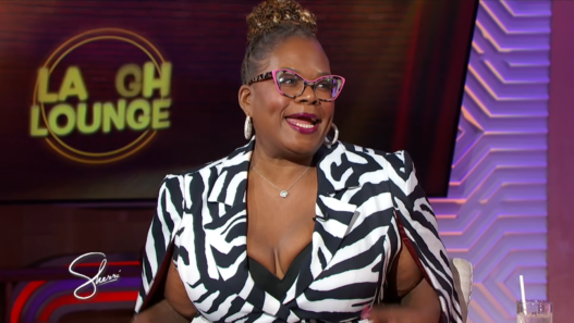 Woman guest wearing a black-and-white zebra print blazer with pink glasses and large hoops on a TV talk show set.