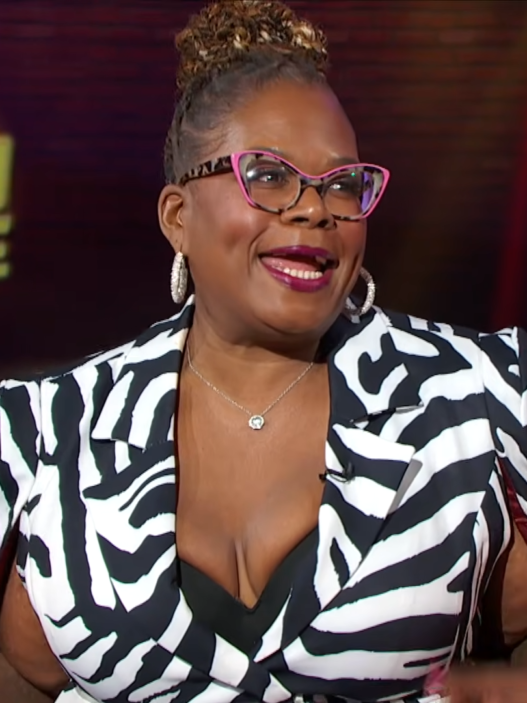 Woman guest wearing a black-and-white zebra print blazer with pink glasses and large hoops on a TV talk show set.