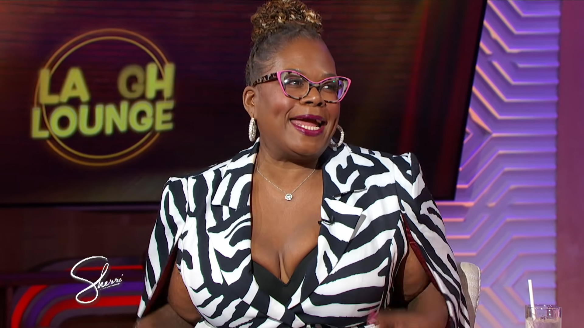 Woman guest wearing a black-and-white zebra print blazer with pink glasses and large hoops on a TV talk show set.