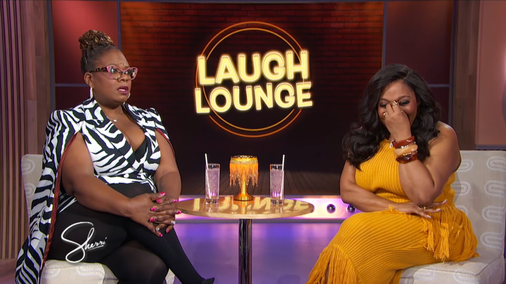 Two women sit on a Laugh Lounge set with a glowing neon sign behind them and drinks on the table in front of them.