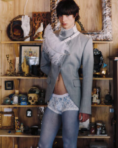 Young model in a light gray suit with white feather accessory, standing in a cluttered shelves backdrop of knickknacks and decor.