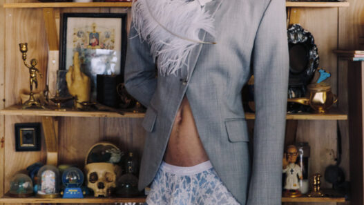 Young model in a light gray suit with white feather accessory, standing in a cluttered shelves backdrop of knickknacks and decor.