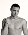 Bare-chested man with short dark hair wearing a pearl necklace and bracelets, gazing at the camera with tattoos on his chest and arms.