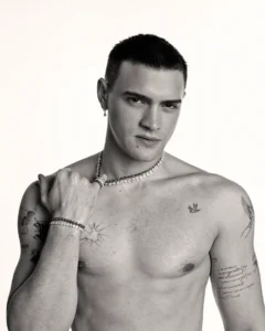 Bare-chested man with short dark hair wearing a pearl necklace and bracelets, gazing at the camera with tattoos on his chest and arms.