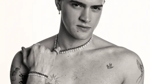 Bare-chested man with short dark hair wearing a pearl necklace and bracelets, gazing at the camera with tattoos on his chest and arms.