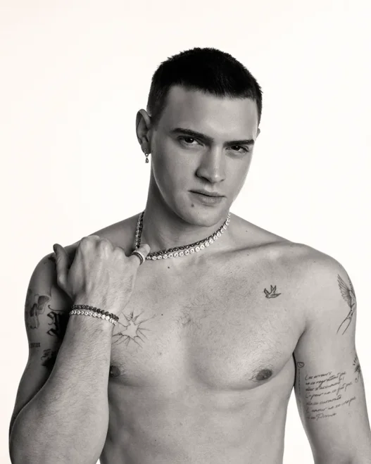 Bare-chested man with short dark hair wearing a pearl necklace and bracelets, gazing at the camera with tattoos on his chest and arms.