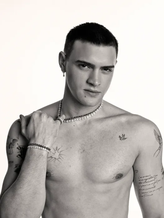 Bare-chested man with short dark hair wearing a pearl necklace and bracelets, gazing at the camera with tattoos on his chest and arms.