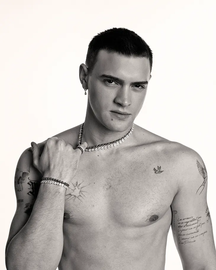 Bare-chested man with short dark hair wearing a pearl necklace and bracelets, gazing at the camera with tattoos on his chest and arms.