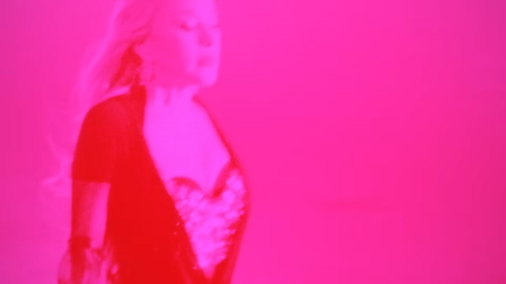 Woman in lingerie standing in a pink-lit scene, facing left.