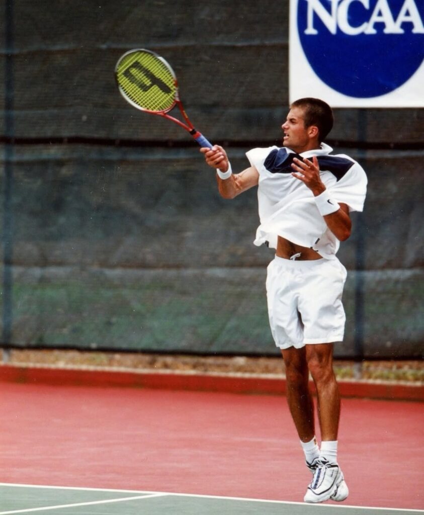 Tennis player in a white outfit mid-air forehand on a red-clay court.