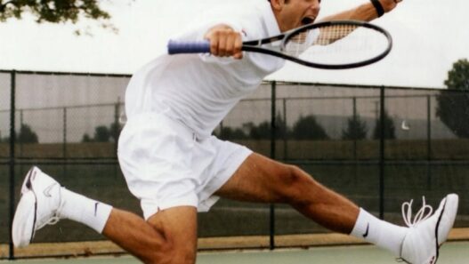Male tennis player in white outfit leaping to hit a volley on an outdoor court.