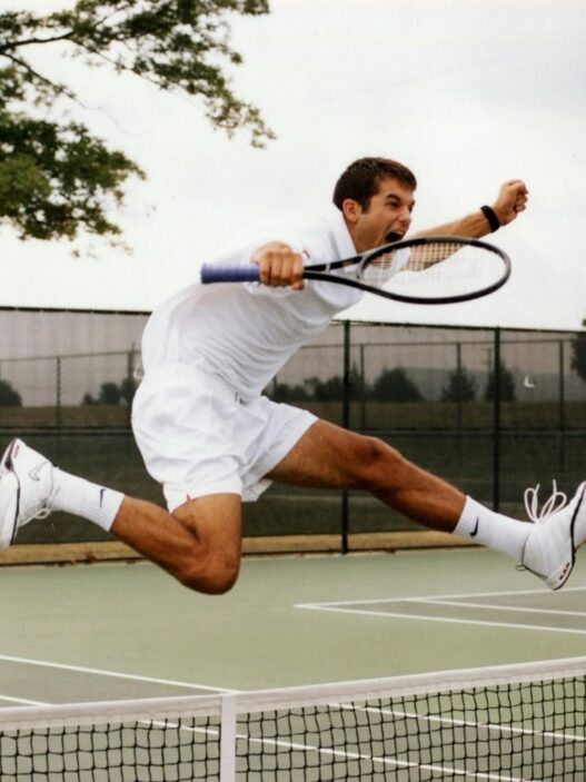 Male tennis player in white outfit leaping to hit a volley on an outdoor court.