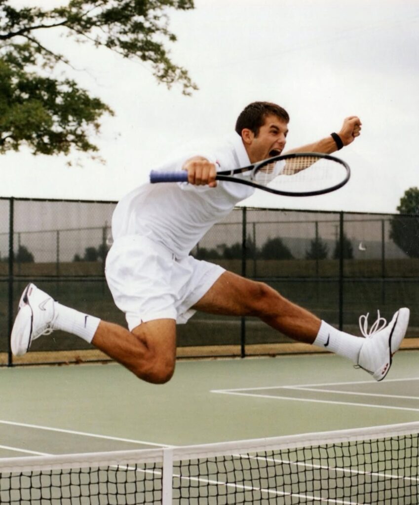 Male tennis player in white outfit leaping to hit a volley on an outdoor court.