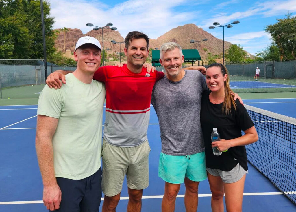 Four friends on a blue tennis court with mountains in the background, smiling with arms around each other.