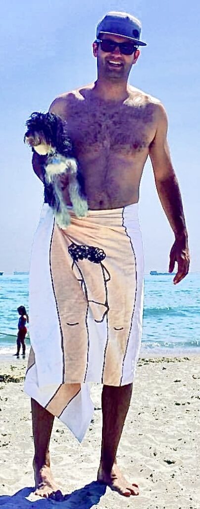 Shirtless man in a cap and sunglasses on a sandy beach, holding a small dog in his left arm with the ocean in the background.