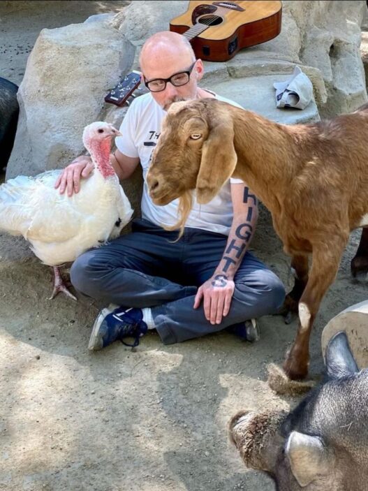 Man with glasses sits cross-legged on dirt, petting a white turkey while a brown goat stands nearby in a rocky outdoor setting; a guitar rests on the rocks behind them.