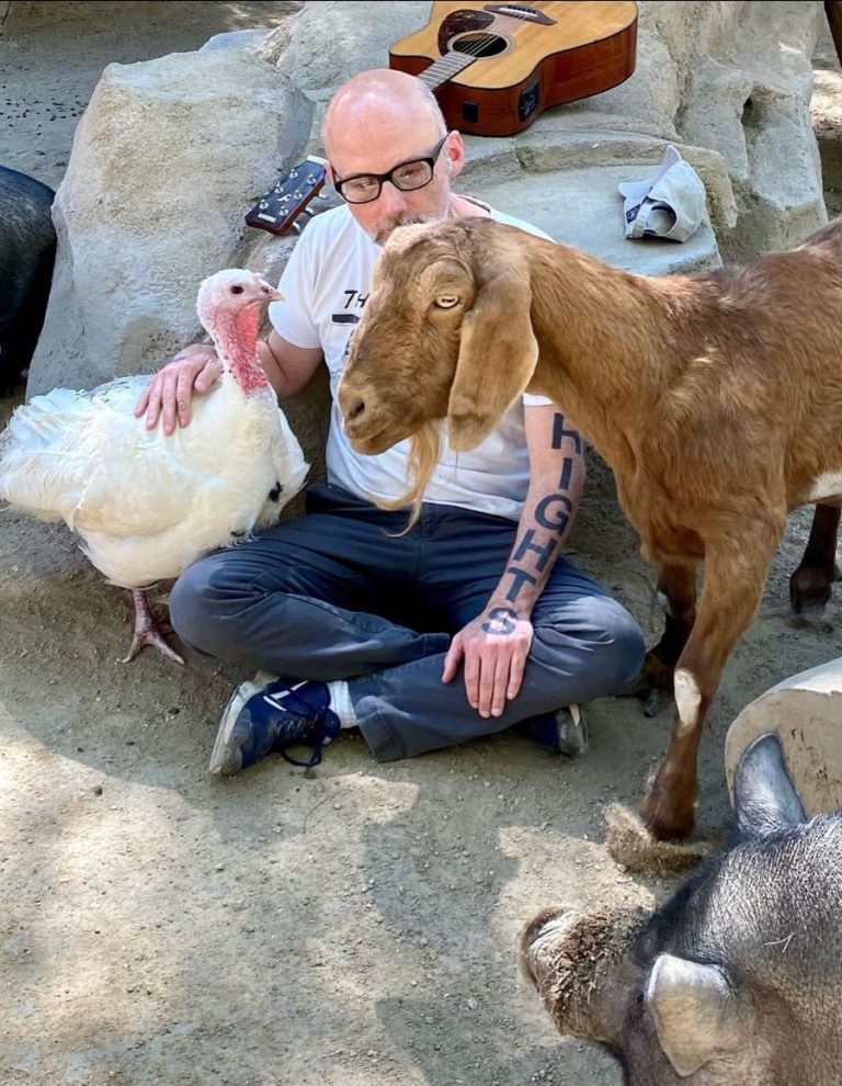 Man with glasses sits cross-legged on dirt, petting a white turkey while a brown goat stands nearby in a rocky outdoor setting; a guitar rests on the rocks behind them.