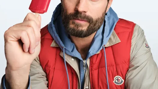Bearded man in beige and red jacket holding a red popsicle and looking at the camera.