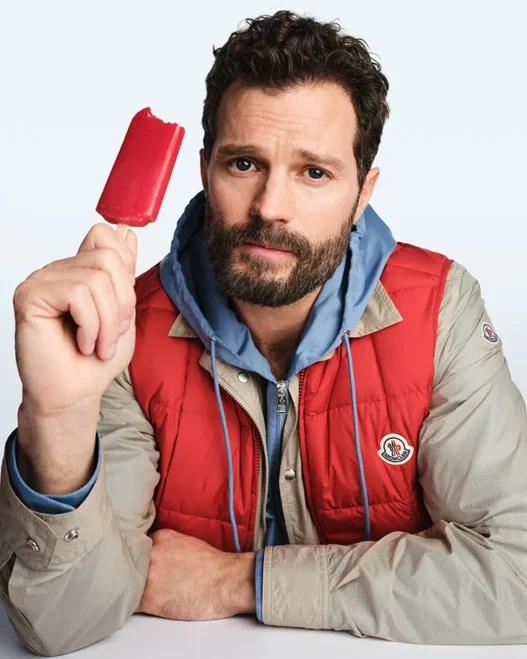 Bearded man in beige and red jacket holding a red popsicle and looking at the camera.