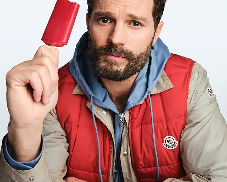 Bearded man in beige and red jacket holding a red popsicle and looking at the camera.
