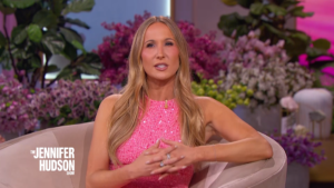 A woman in a pink sequined dress sits on a beige sofa, speaking on a talk-show set with purple and pink flowers in the background, logo visible on the chair edge.