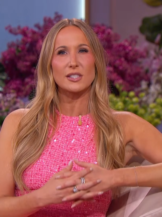 A woman in a pink sequined dress sits on a beige sofa, speaking on a talk-show set with purple and pink flowers in the background, logo visible on the chair edge.