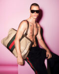 Shirtless male model with dark sunglasses sits against a pink backdrop, holding a large Gucci monogram bag over his shoulder; visible chest tattoos.