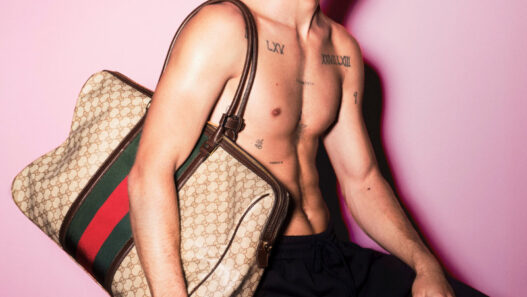 Shirtless male model with dark sunglasses sits against a pink backdrop, holding a large Gucci monogram bag over his shoulder; visible chest tattoos.