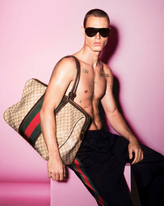 Shirtless male model with dark sunglasses sits against a pink backdrop, holding a large Gucci monogram bag over his shoulder; visible chest tattoos.
