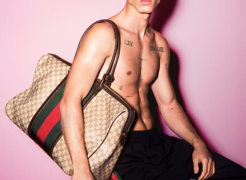 Shirtless male model with dark sunglasses sits against a pink backdrop, holding a large Gucci monogram bag over his shoulder; visible chest tattoos.