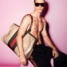 Shirtless male model with dark sunglasses sits against a pink backdrop, holding a large Gucci monogram bag over his shoulder; visible chest tattoos.