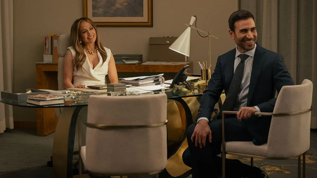 Two professionally dressed colleagues sit at a glass desk in a modern office, smiling at each other.