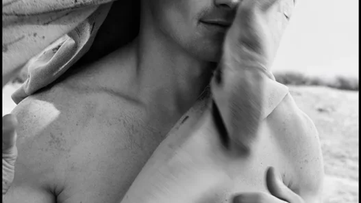 Black-and-white photo of a shirtless man outdoors with a towel draped over his head, looking at the camera.