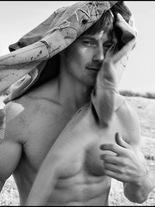 Black-and-white photo of a shirtless man outdoors with a towel draped over his head, looking at the camera.
