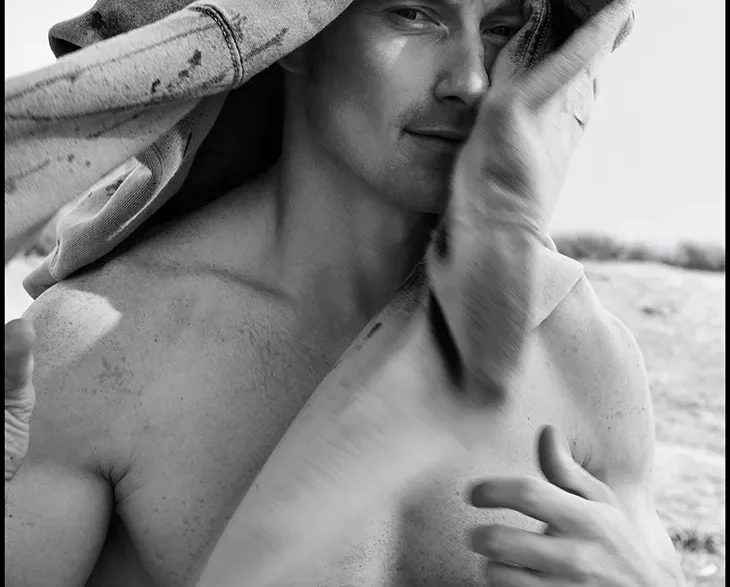 Black-and-white photo of a shirtless man outdoors with a towel draped over his head, looking at the camera.