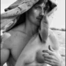 Black-and-white photo of a shirtless man outdoors with a towel draped over his head, looking at the camera.