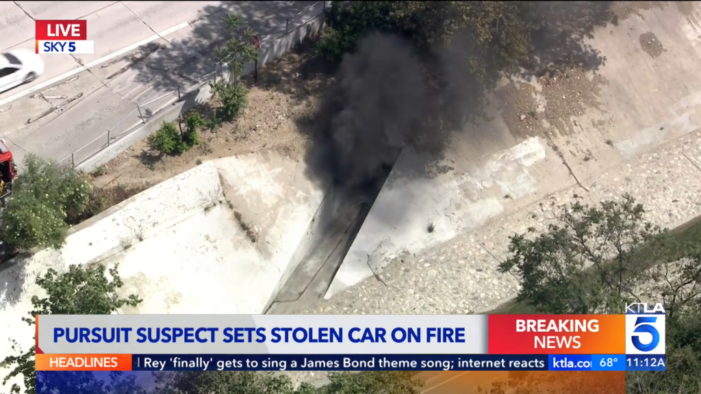 Aerial view of a car on fire in a concrete canal beside a road, black smoke billowing upward as firefighters or equipment are nearby; breaking news banner at the bottom.