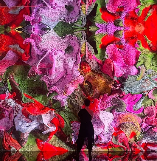 Person silhouetted in front of a large kaleidoscopic mural with pink, red, purple, and green floral patterns on a wall inside an installation.