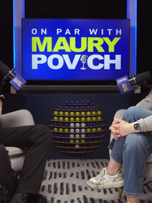 Two podcast hosts sit in armchairs facing each other, smiling and talking, with a blue sign reading 'ON PAR WITH MAURY POVICH' behind them and a rack of golf balls between.