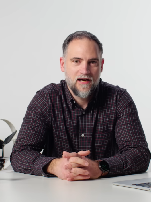 Man in plaid shirt sits at a desk with a disassembled drone and a laptop, speaking to camera in a studio setup.