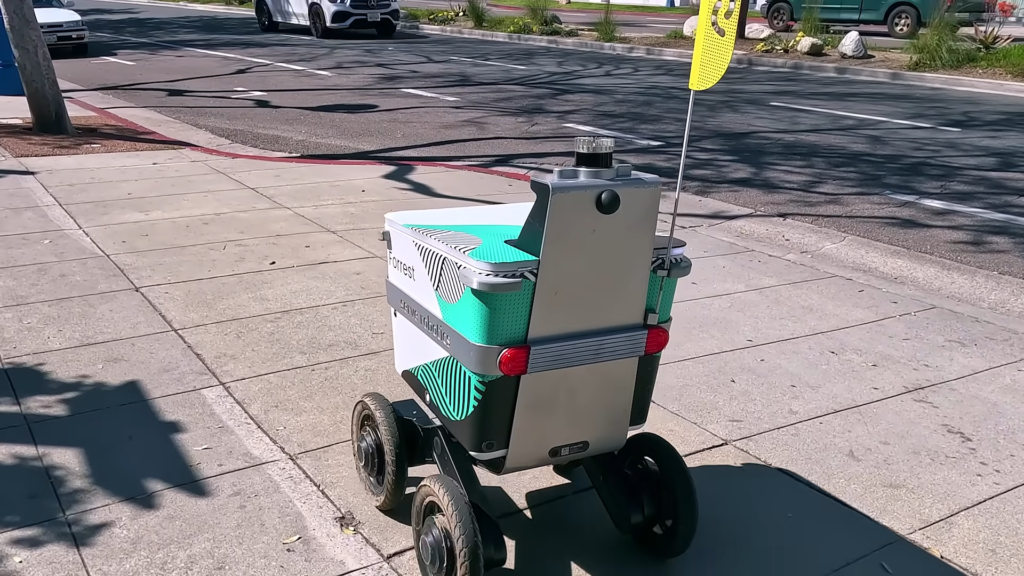 A portable, wheeled cooler cart on a sidewalk with teal accents and a yellow flag nearby, in a sunny urban area.