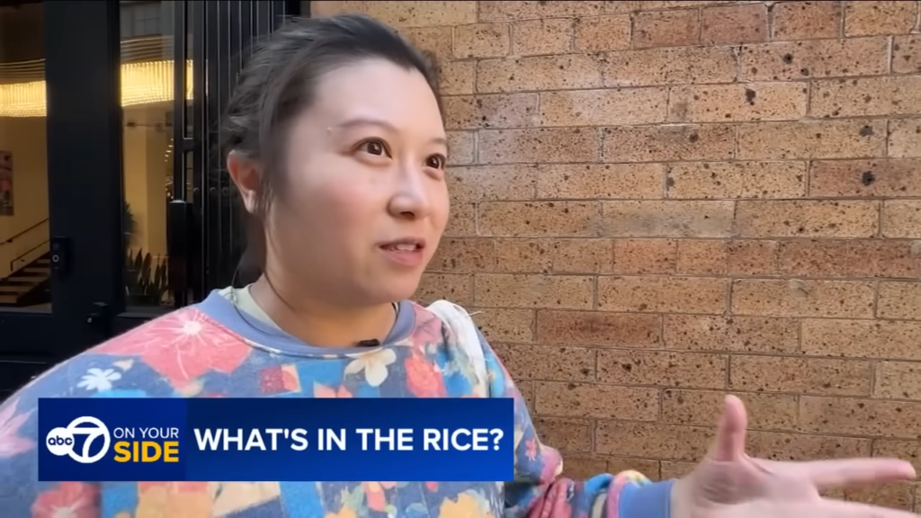 Woman being interviewed outdoors, gesturing with her hand, with a blue caption bar reading 'WHAT'S IN THE RICE?' and brick wall backdrop.