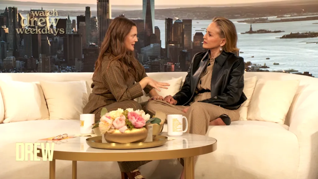 Two women sit on a white couch in a talk-show studio, facing each other as they converse, with a city skyline backdrop and a floral arrangement on a coffee table in front of them.